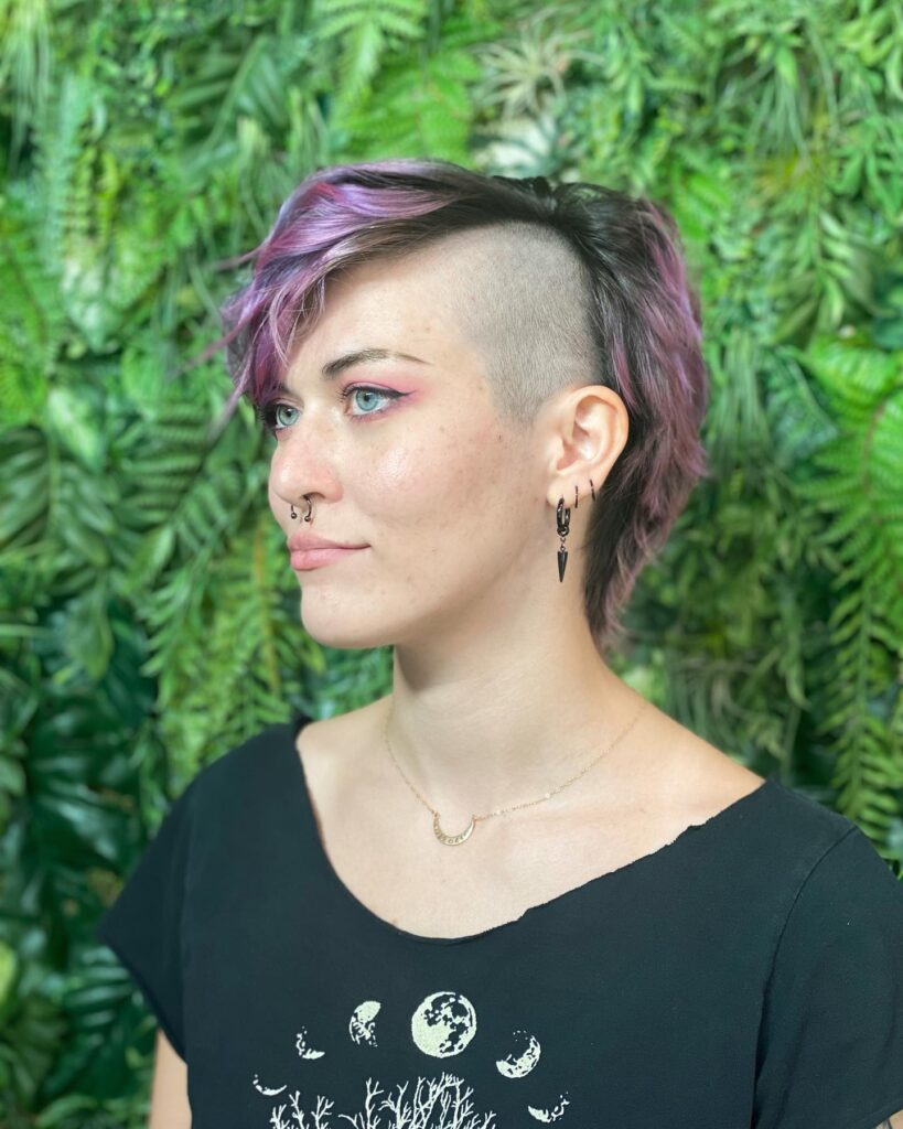 A person with a short, smoky lavender hairstyle featuring a clean side shave and a textured side-swept fringe.