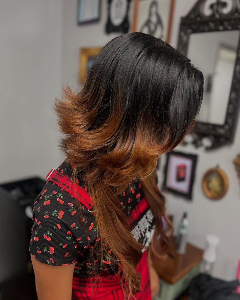 A woman with long hair featuring black roots and copper brown ends styled in flared, shaggy layers.