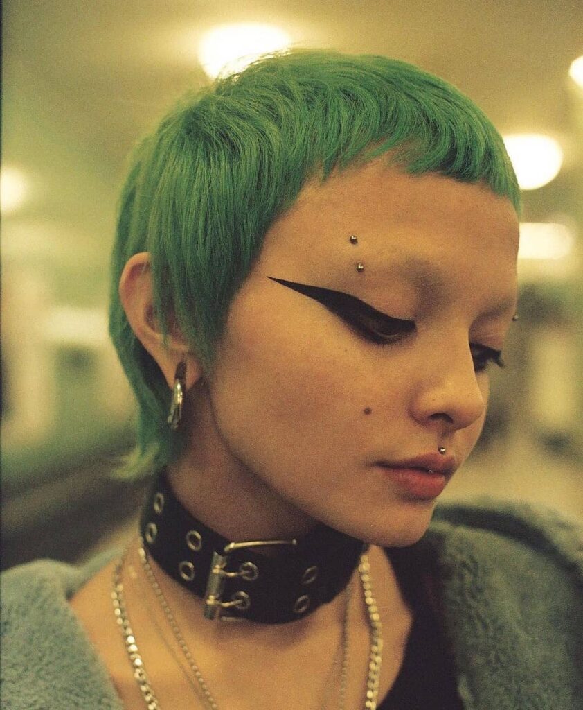 A person with a short seafoam green micro mullet featuring textured baby bangs and shaggy layers at the neck.