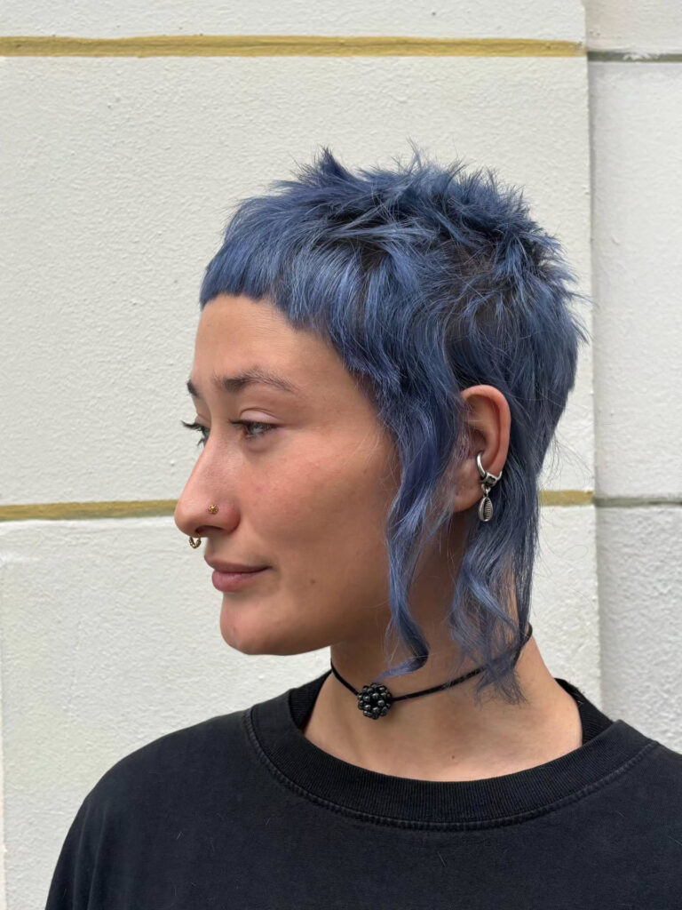 A woman with a short, dusty blue pixie mullet featuring a micro fringe and long wispy curls at the side.