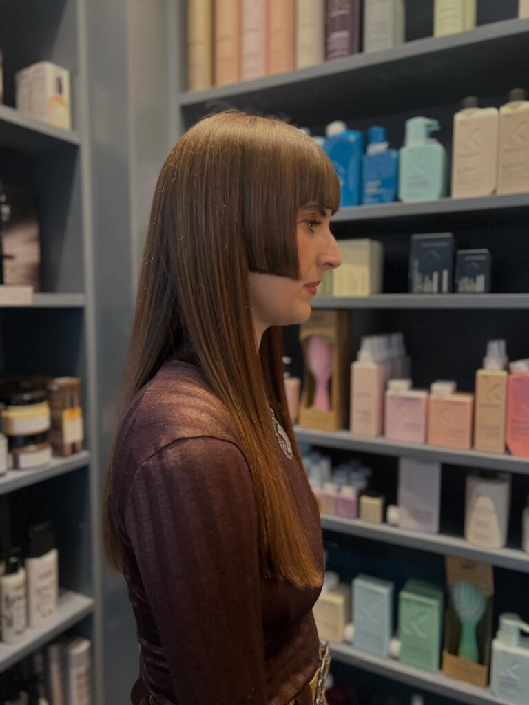 A woman viewed from the side with long, straight brown hair featuring a blunt fringe and sharp geometric hime-style layers at the cheekbones.