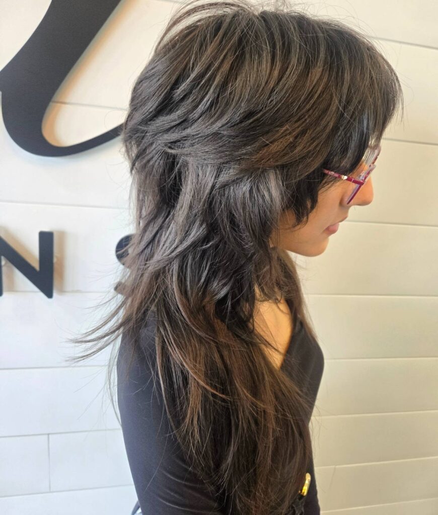 A woman viewed from the side with a long, heavily layered wolf cut in a dark ash brunette shade, featuring a voluminous crown and wispy ends.