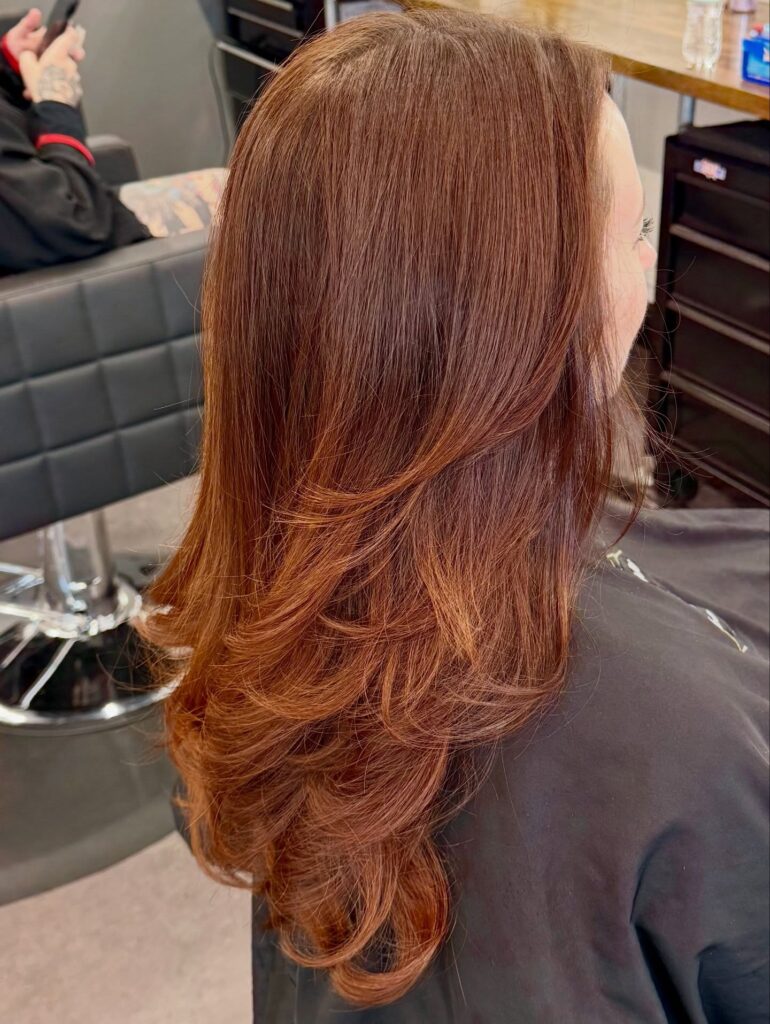 A woman with long copper red hair featuring soft feathered layers and bouncy curled ends.