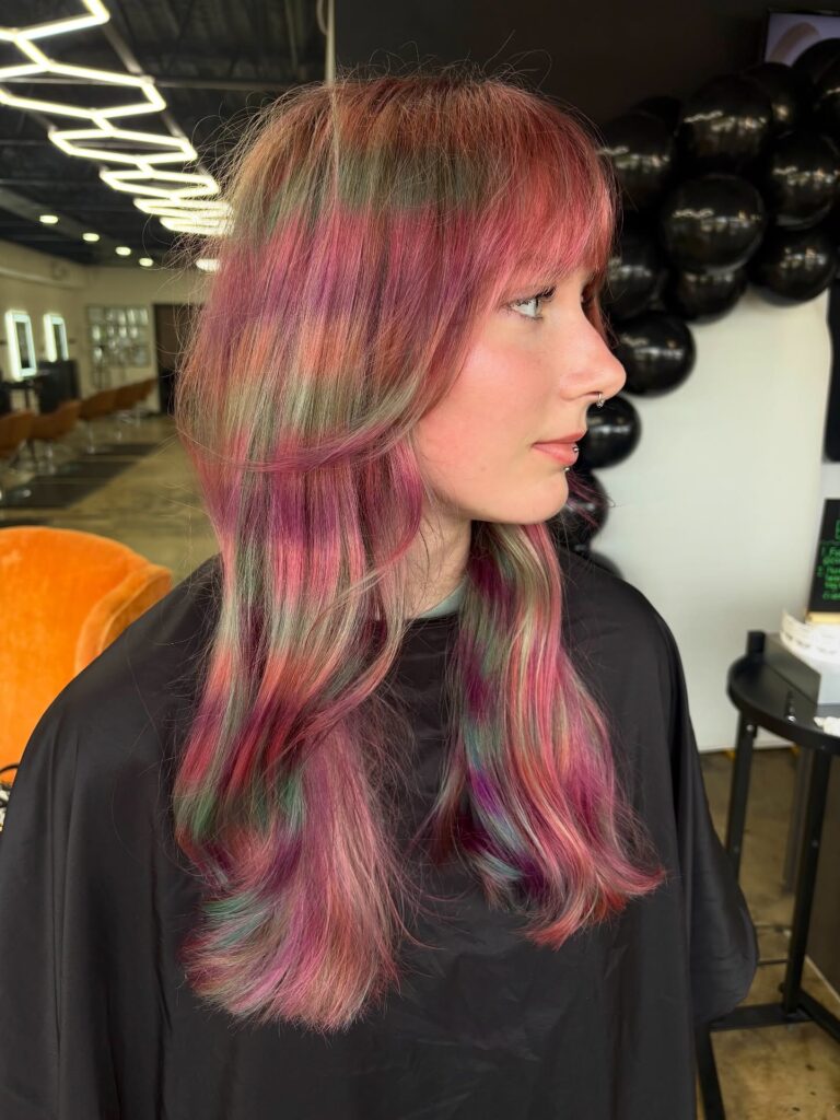 A woman viewed from the side with long layered hair featuring horizontal bands of pink, purple, and green colors and soft bangs.