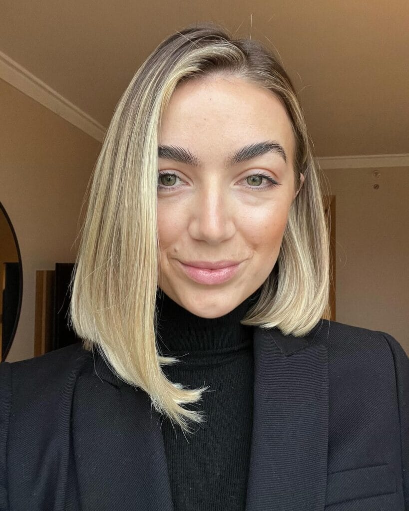 A woman with a straight sandy blonde medium lob featuring a blunt cut and bright, face-framing highlights.