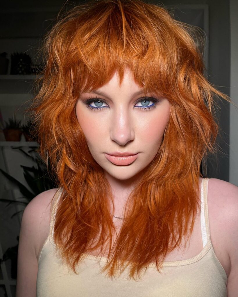 A woman with a mid-length bright apricot copper shag haircut and thick choppy bangs.
