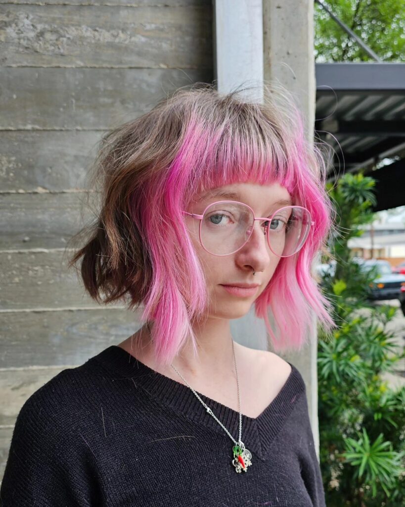 A woman with a short brown bob featuring a vibrant pink money piece and matching pink bangs.