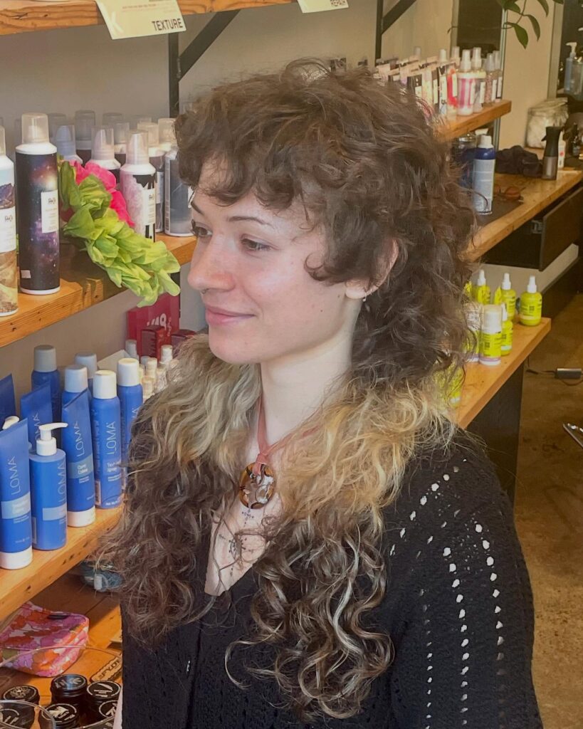 A woman with long curly hair featuring dark brown roots and bright blonde ends styled in a shaggy, layered cut.