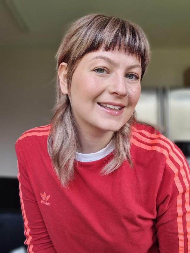 A woman with a mushroom brown wolf cut featuring ash-blonde highlights and a very short, straight fringe.