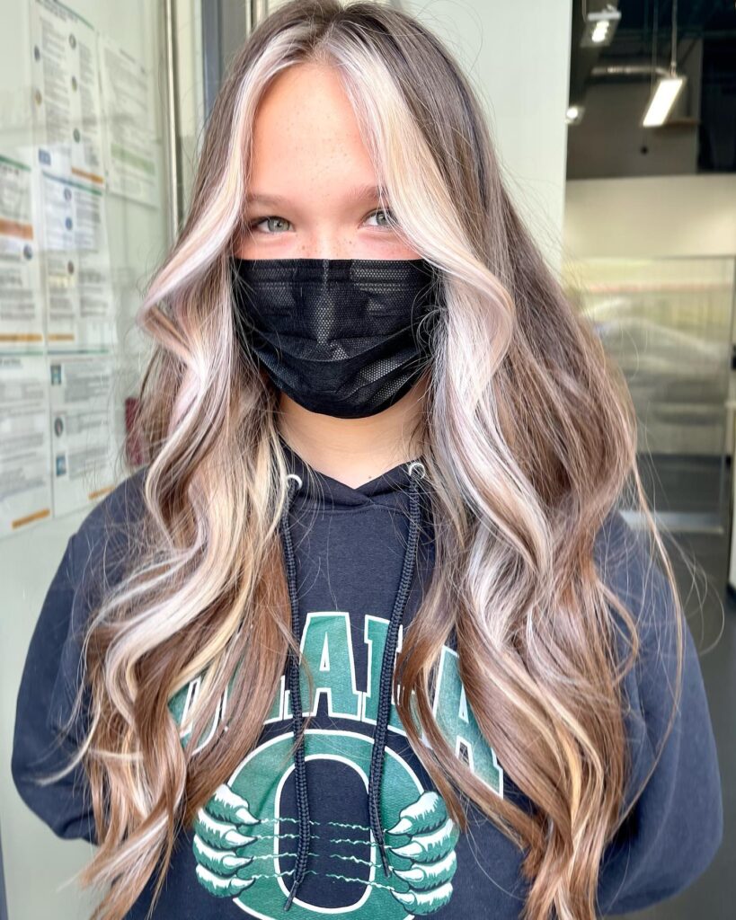 A woman with long, wavy, brown hair featuring bold, creamy blonde money piece highlights and face-framing layers.