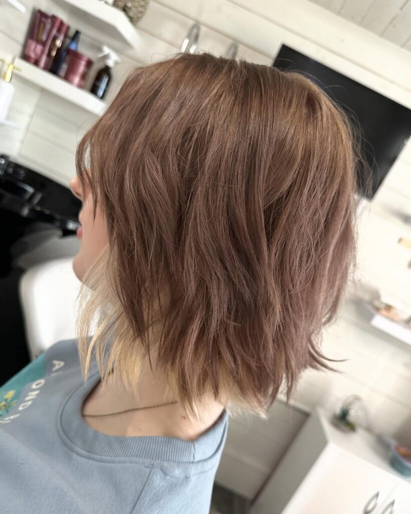 A woman with a textured mauve brown shag haircut featuring a pale blonde peekaboo highlight layer underneath.