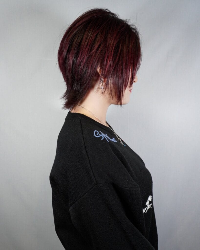 A side profile photograph of a woman wearing a modern short mullet with choppy layers and long wispy hair in the back, colored in a deep burgundy hue.