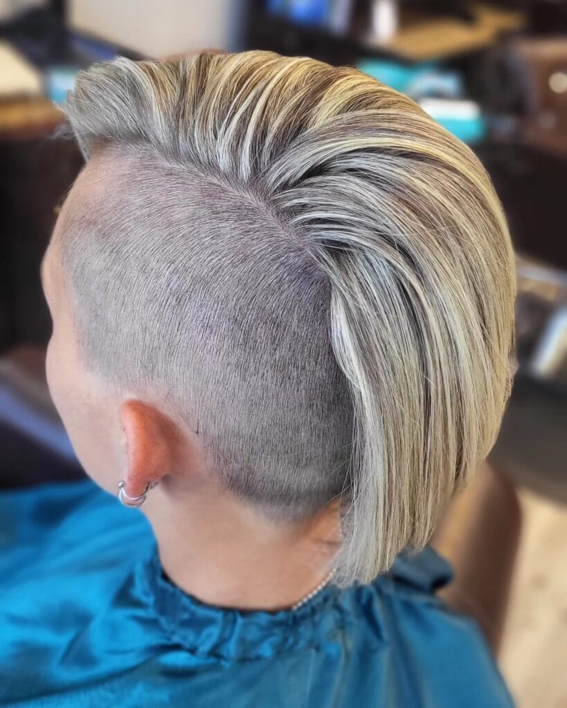 A side and back view of a woman with a short ash blonde mohawk hairstyle featuring shaved sides and long swept back hair on top.