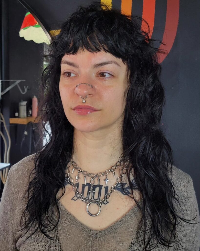 A woman with a long, black, shaggy wolf cut featuring a choppy fringe and textured wavy layers.