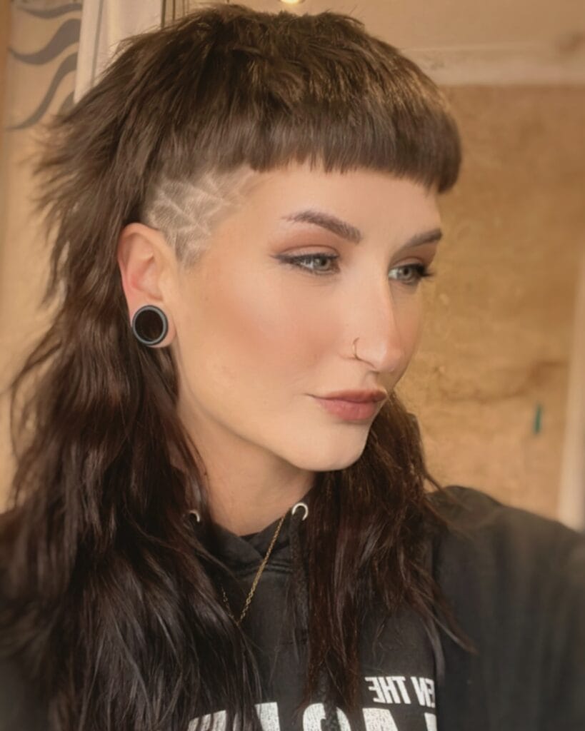A woman with a brown mullet featuring a blunt micro fringe and a shaved side undercut with a geometric design.