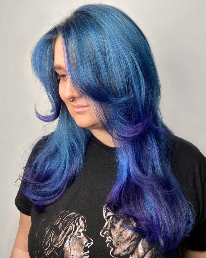 A woman with long hair featuring a blue to purple ombre color and soft feathered layers that flip outward at the ends.