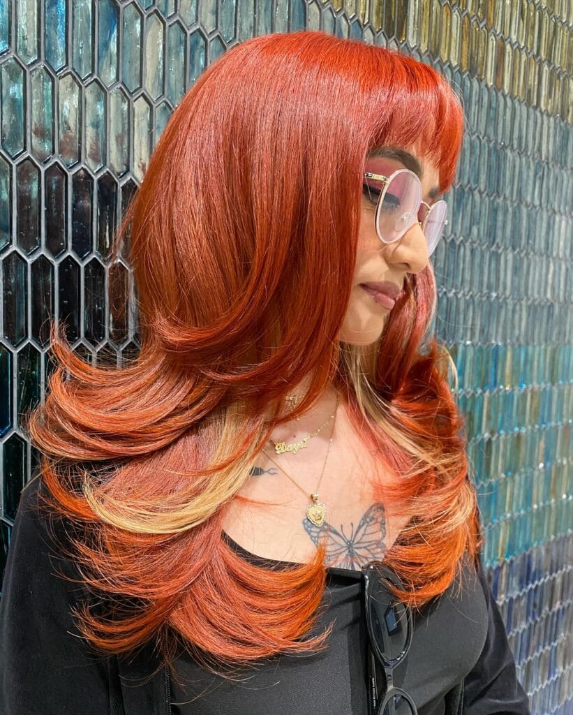 A woman with long copper hair featuring blonde highlights and voluminous tiered layers that flip outward.