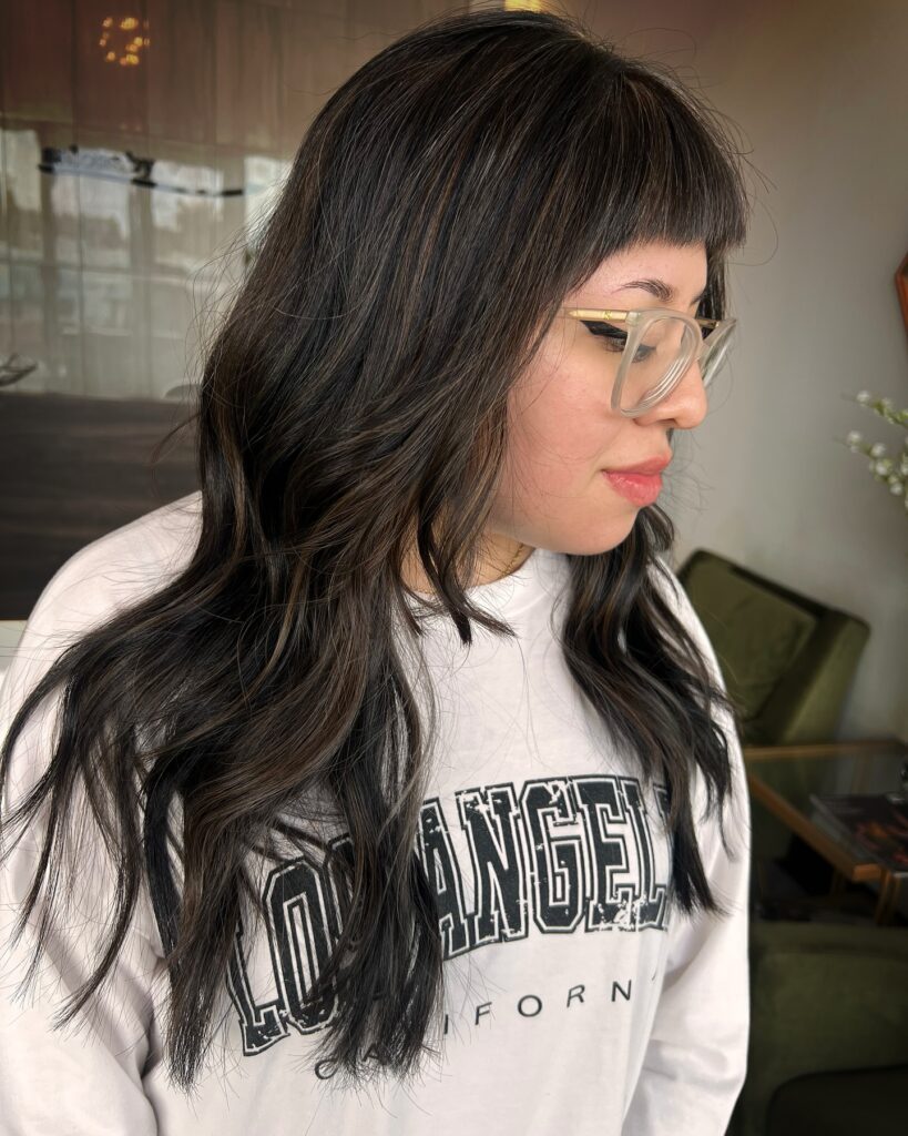 A woman with long, dark, wavy hair featuring subtle brown highlights and short, blunt bangs.