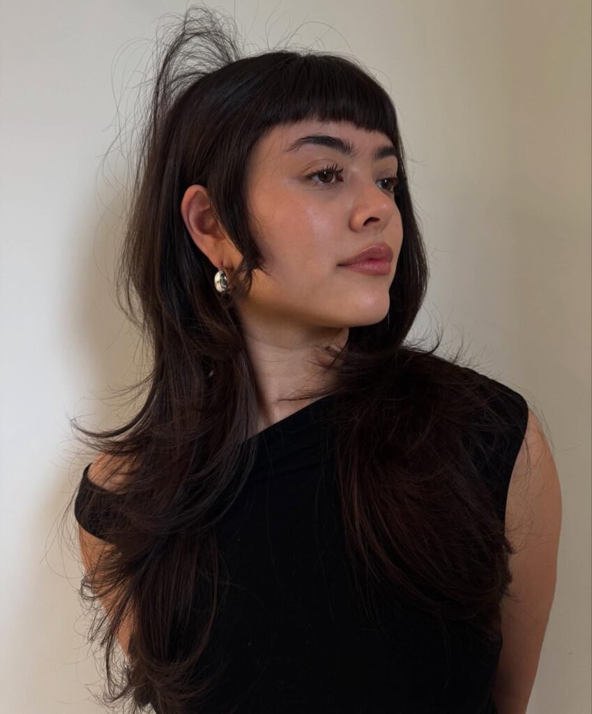 A woman with long, layered dark brown hair and short arched bangs that frame her forehead.