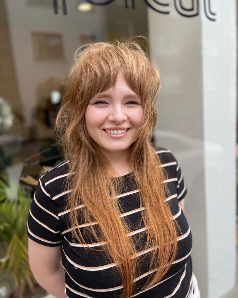 A smiling woman with long copper-colored hair in a shaggy wolf cut with feathered bangs.