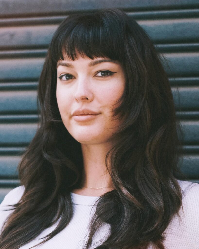 A woman with long, wavy dark brown hair and thick, curved bangs that frame her face.
