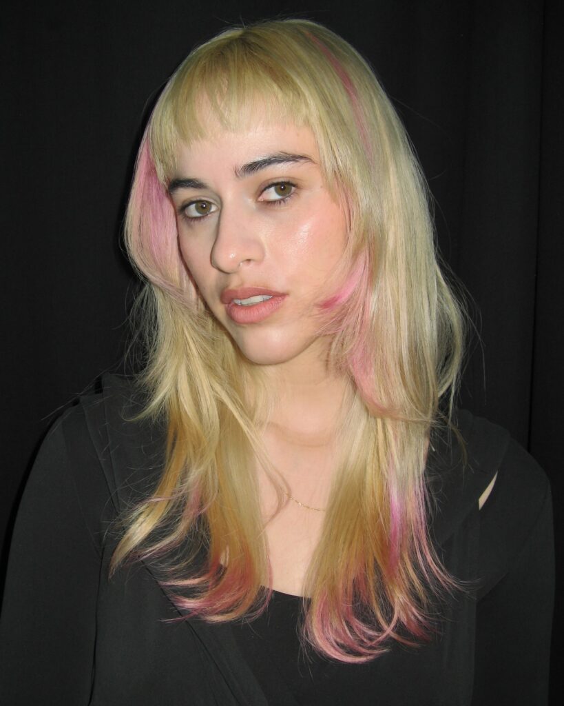A woman with long blonde hair featuring pink highlights and sharp chin-length side layers with short bangs.