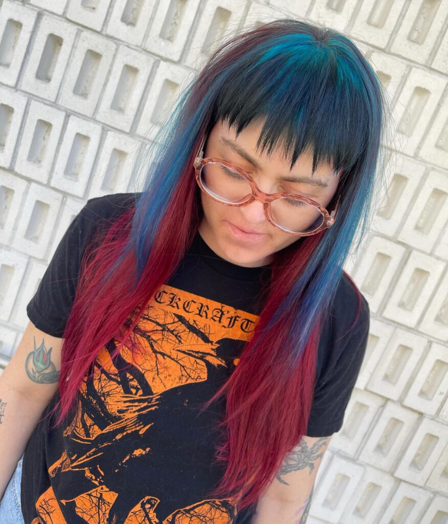 A woman with long straight hair featuring burgundy and blue color-blocking and short jagged bangs.