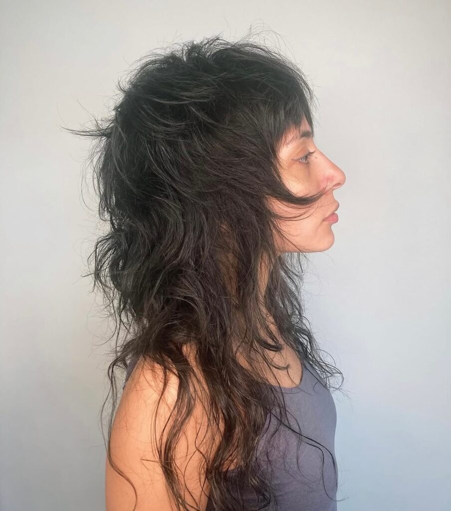A side profile of a woman with a long dark shaggy mullet featuring voluminous crown layers and feathered bangs.