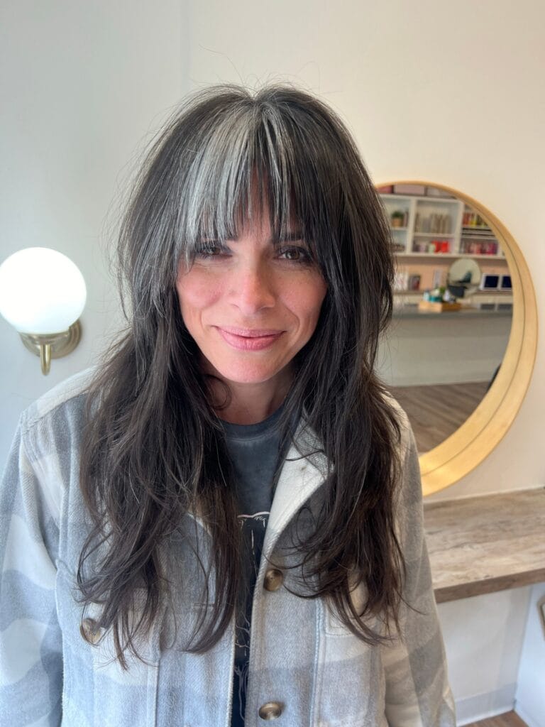 A woman with long dark layered hair featuring a silver money piece in her bangs.