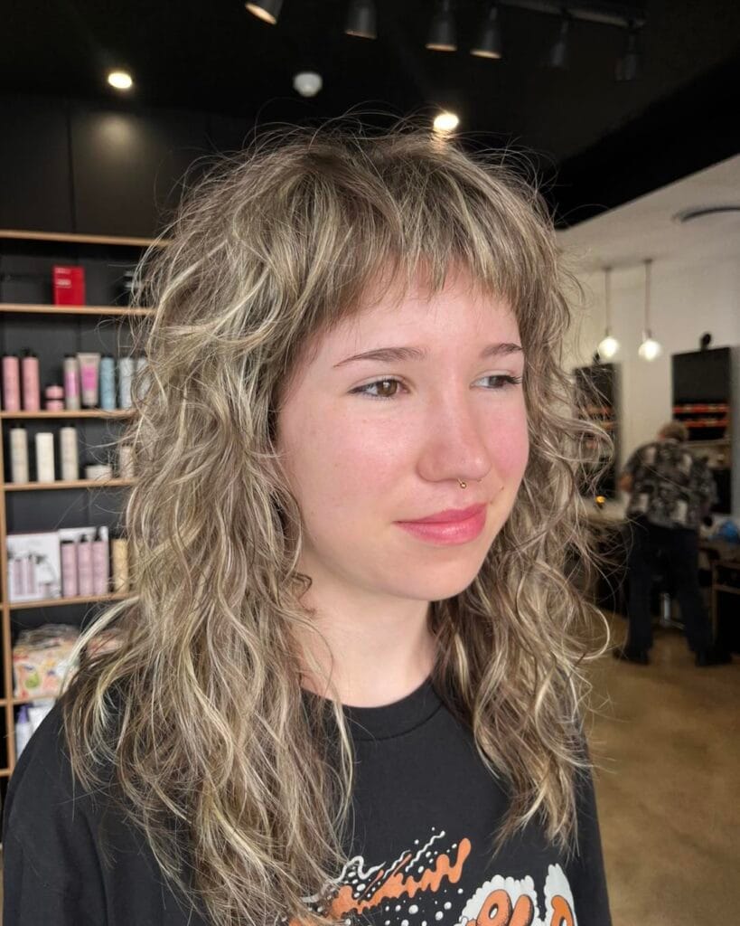 A woman with long curly ash-blonde hair and shaggy bangs smiling in a salon.