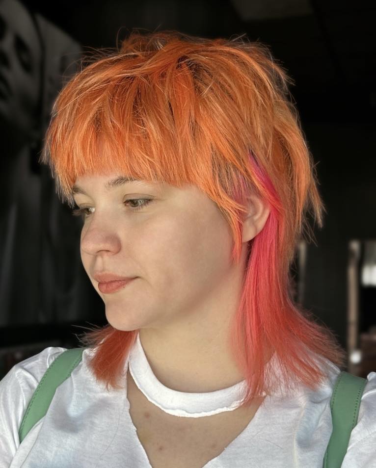 A woman with a neon orange and pink layered wolf cut with shaggy bangs.