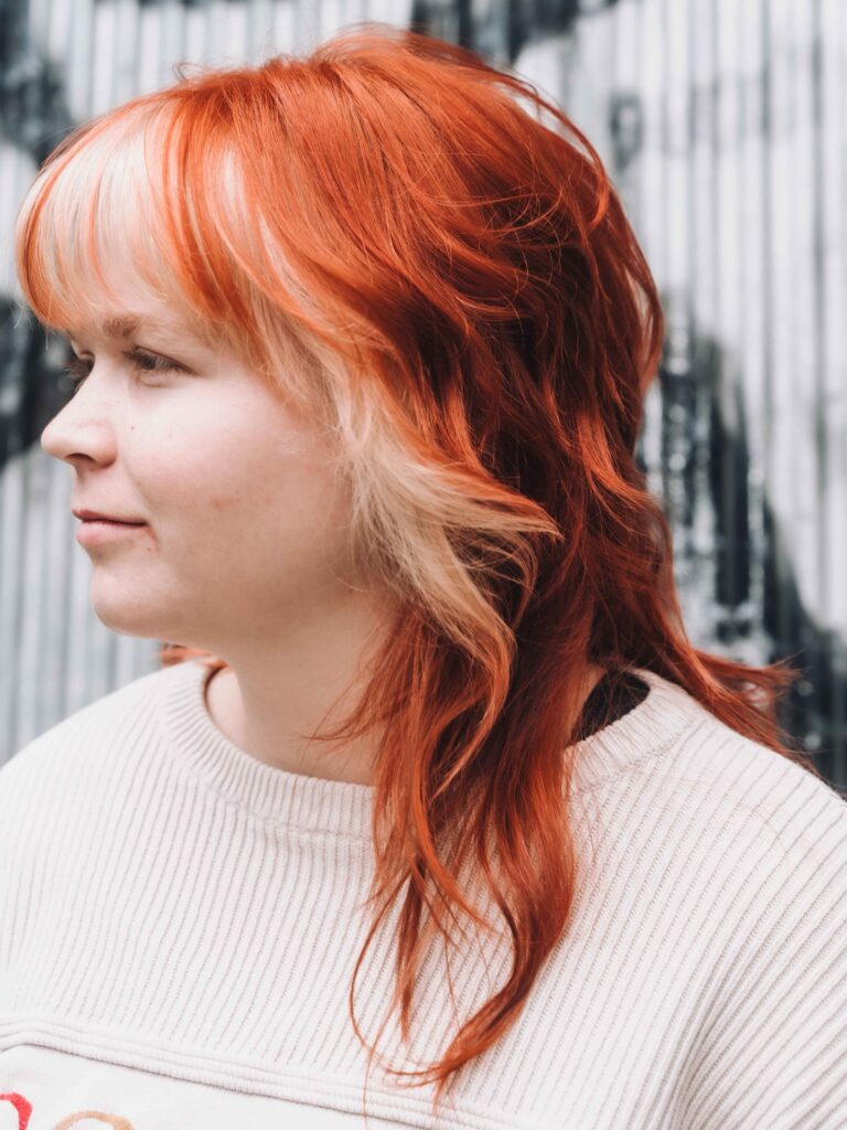 A woman with a layered copper red shag haircut and bright blonde money-piece highlights in her bangs.