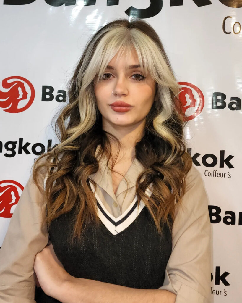 A woman with long, wavy brown hair and full platinum blonde bangs and face-framing highlights.