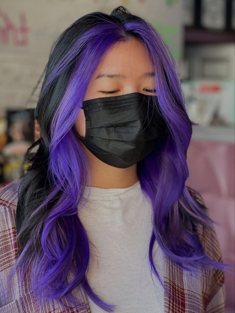 A woman with long, wavy black hair featuring a vibrant electric purple money piece framing her face.