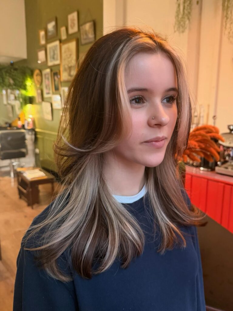 A woman with long, layered brunette hair and bright, sandy blonde face-framing highlights styled with feathered ends.