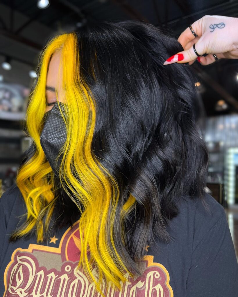 A woman with medium-length wavy black hair featuring a thick and vibrant neon yellow money piece framing her face.