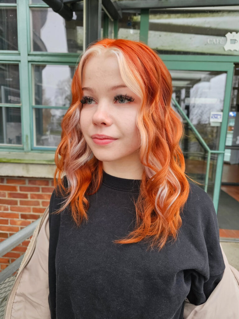 A woman with shoulder-length wavy orange hair and a creamy pearl-blonde money piece framing her face.