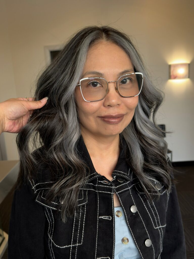 A woman with long, wavy, dark charcoal hair and a bright, icy silver money piece framing her face.