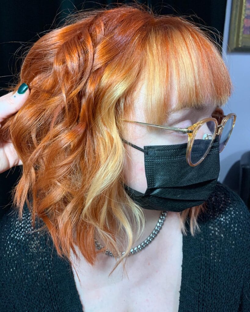 A woman with a copper orange wavy lob featuring light blonde face-framing highlights and a full fringe.