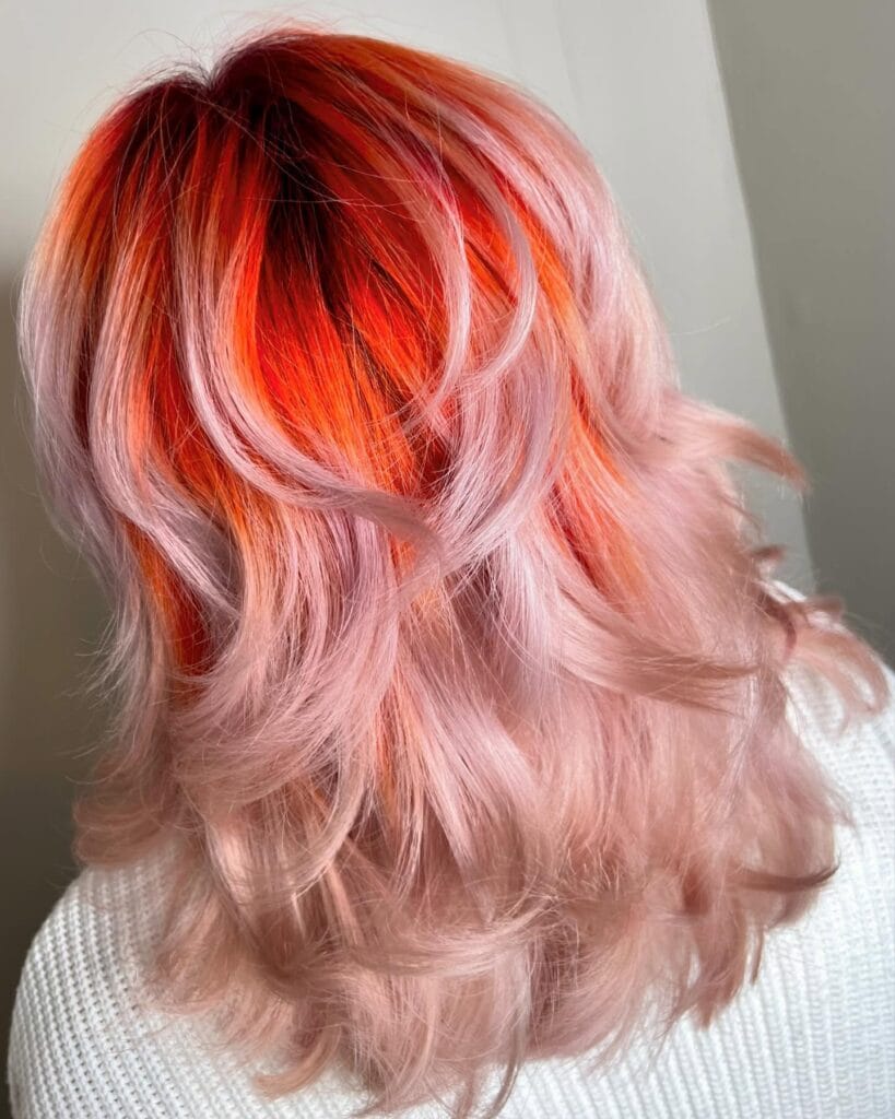 A woman with medium-length layered hair featuring a vibrant coral crown that melts into a soft pastel peach ombre with bouncy curls.