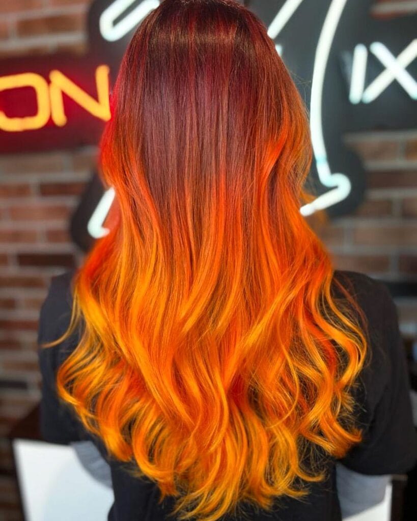 A woman from behind with long wavy hair featuring a crimson-to-bright-orange ombre color.