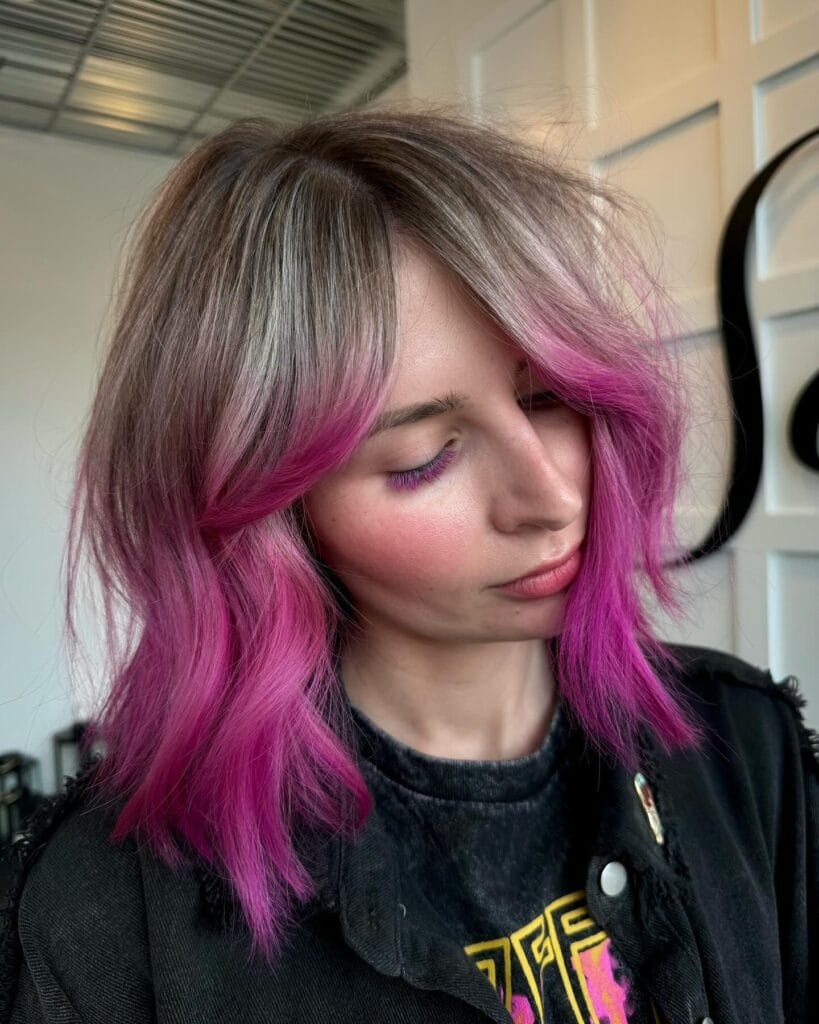A woman with shoulder-length ashy brown hair featuring a vibrant fuchsia pink ombre and face framing curtain bangs.