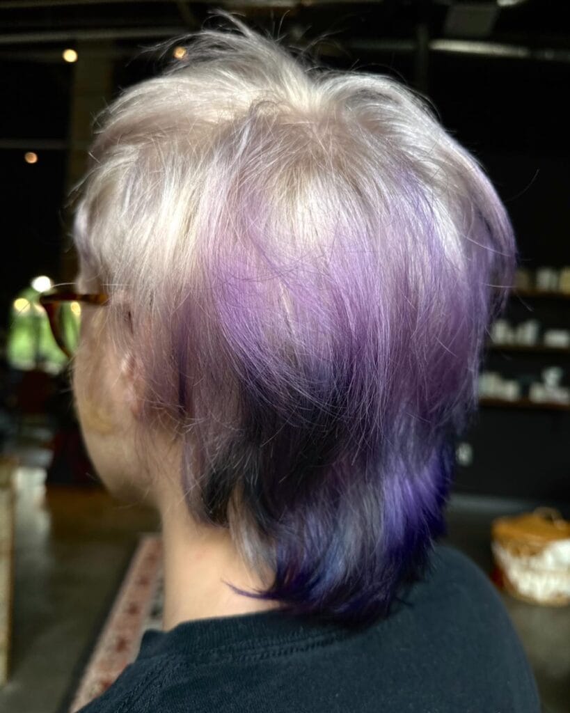 A woman from behind with a short shaggy haircut featuring a platinum blonde crown melting into lavender and indigo purple ombre ends.