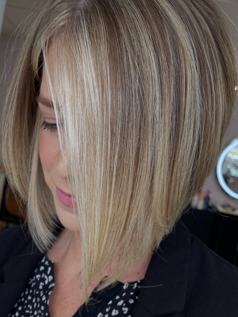 Chin-length angled bob with smooth straight layers and blended blonde highlights, styled sleek with a deep side part that softly frames the face.