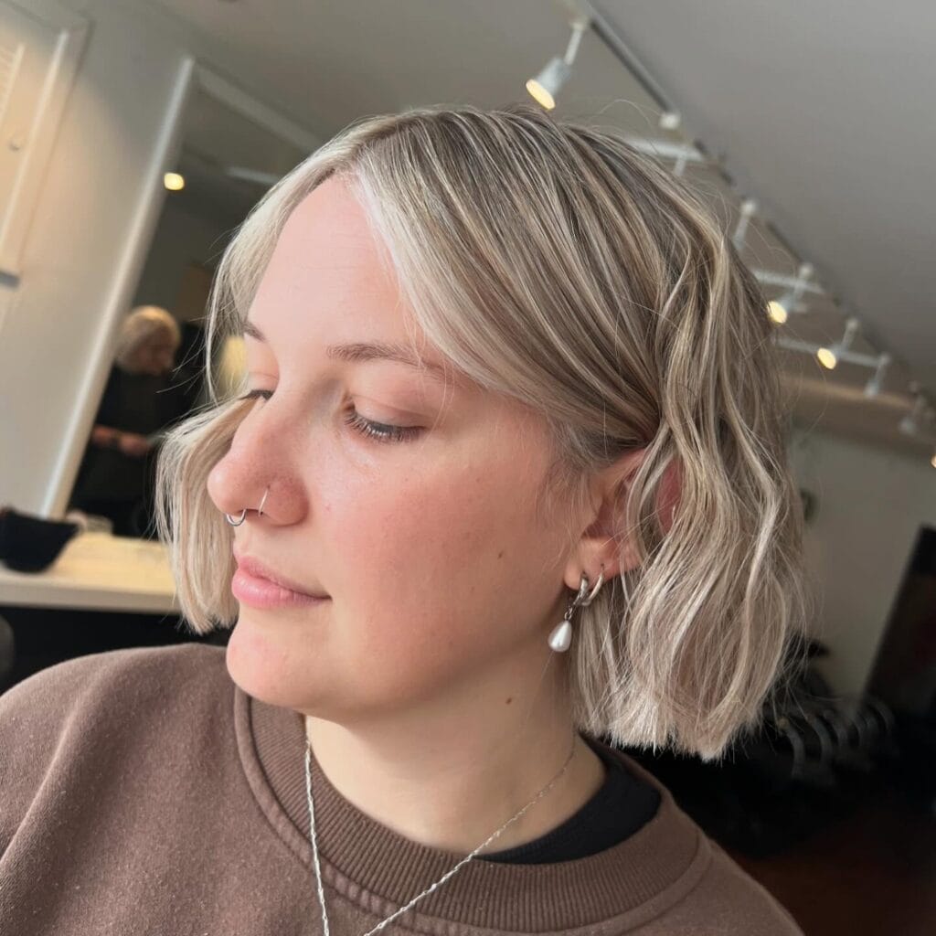 Chin-length wavy blonde bob with a soft center part, subtle darker roots, and loose natural texture for a relaxed, modern look.