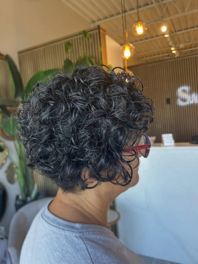 Side view of a short curly pixie bob with dense dark natural curls, softly layered for volume and shape around the back and sides.
