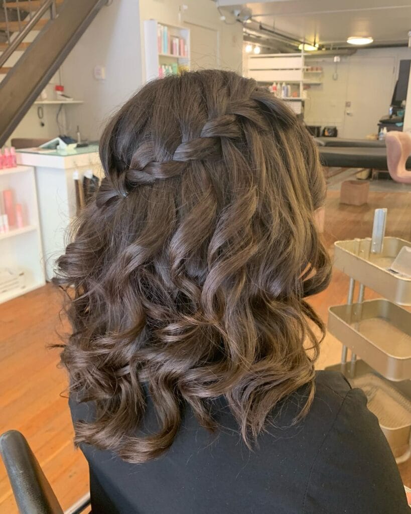 Medium-length dark brown wavy hair styled with a small crown braid across the back, blending into loose, soft curls for a romantic half-up look.