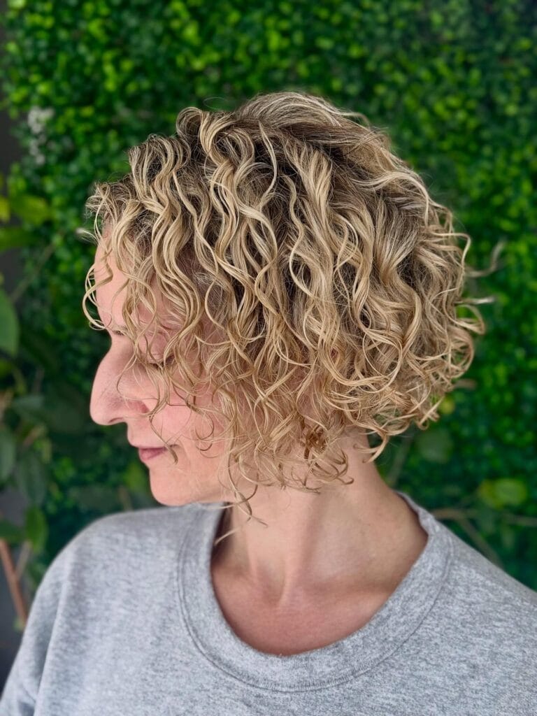 Side profile of a woman with a short, neck-length layered bob featuring natural, tousled curls and blonde highlights that add soft volume and texture.
