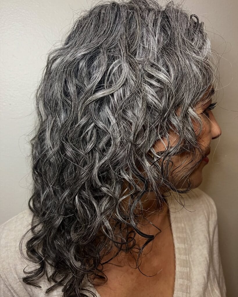 Side view of long, layered naturally curly hair in a silver-gray tone, with soft defined curls cascading past the shoulders and creating full, textured movement.