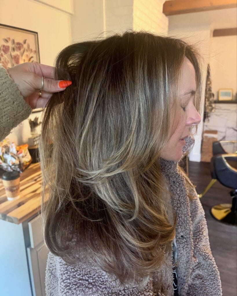 Long layered brunette hair with soft blonde highlights and feathered ends, styled with voluminous layers that add movement and dimension.